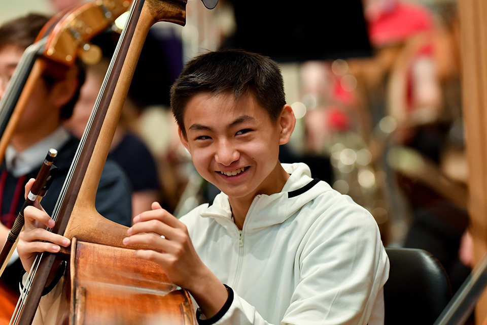 An Asian male RCMJD student, holding his cello, smiling at the camera, sitting in an orchestra.